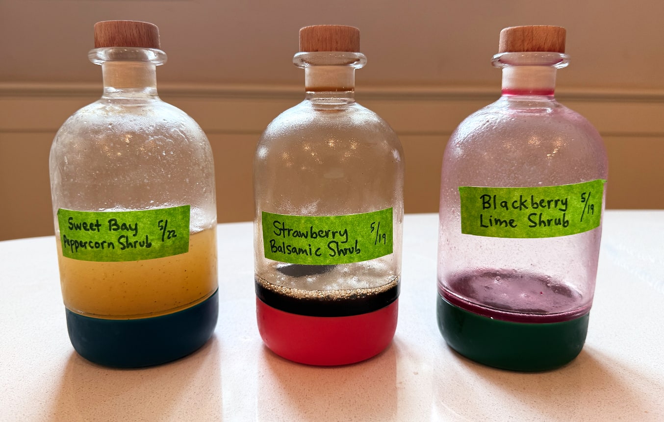Three bottles of shrub syrups labeled “Sweet Bay-Peppercorn Shrub”, “Strawberry Balsamic Shrub”, and “Blackberry-Lime Shrub”